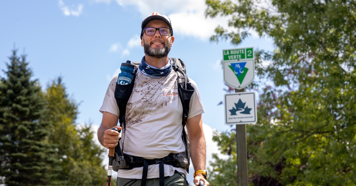 Homme vêtu d'un t-shirt blanc, sac à dos et autres équipements de randonnées avec en arrière plan des arbres et une pancarte de la route Verte n.2
