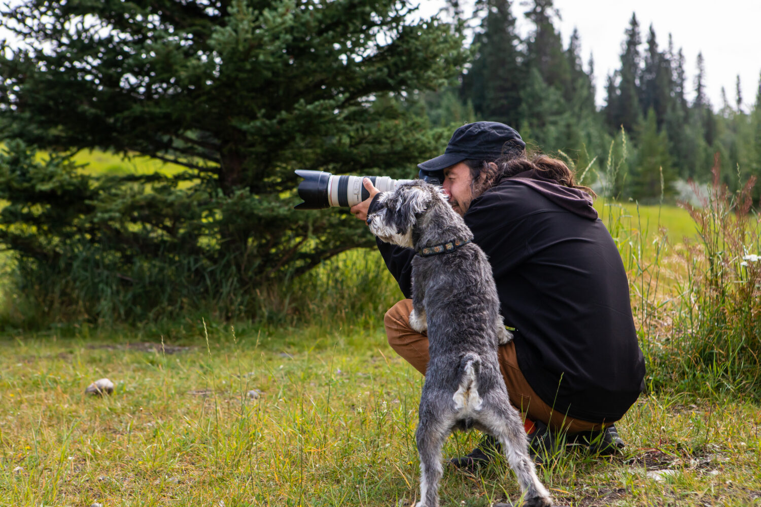 Homme accroupi prenant des photos en nature avec un appareil professionnel et un chien de couleur gris et blanc appuyant ces deux pattes avant sur la cuisse de l'homme.