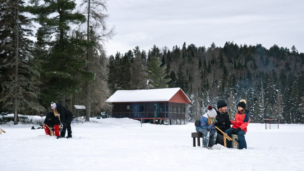 hommes, femmes et enfants pêchant sur la glace avec en arrière plan un chalet rustique