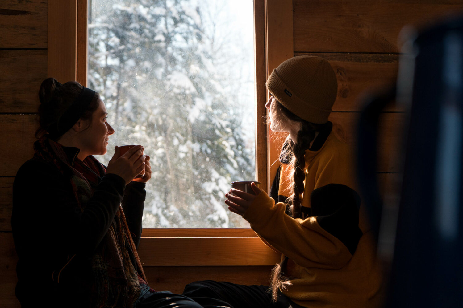 deux femmes qui sont dans un chalet en bois, regarde par la fenêtre en buvant une boisson chaude