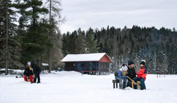 hommes, femmes et enfants pêchant sur la glace avec en arrière plan un chalet rustique